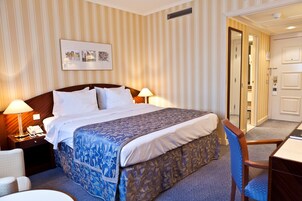 Una camera d'albergo con un letto grande, due lampade sui comodini, un tavolino, una sedia e una porta che dà su un'altra stanza.