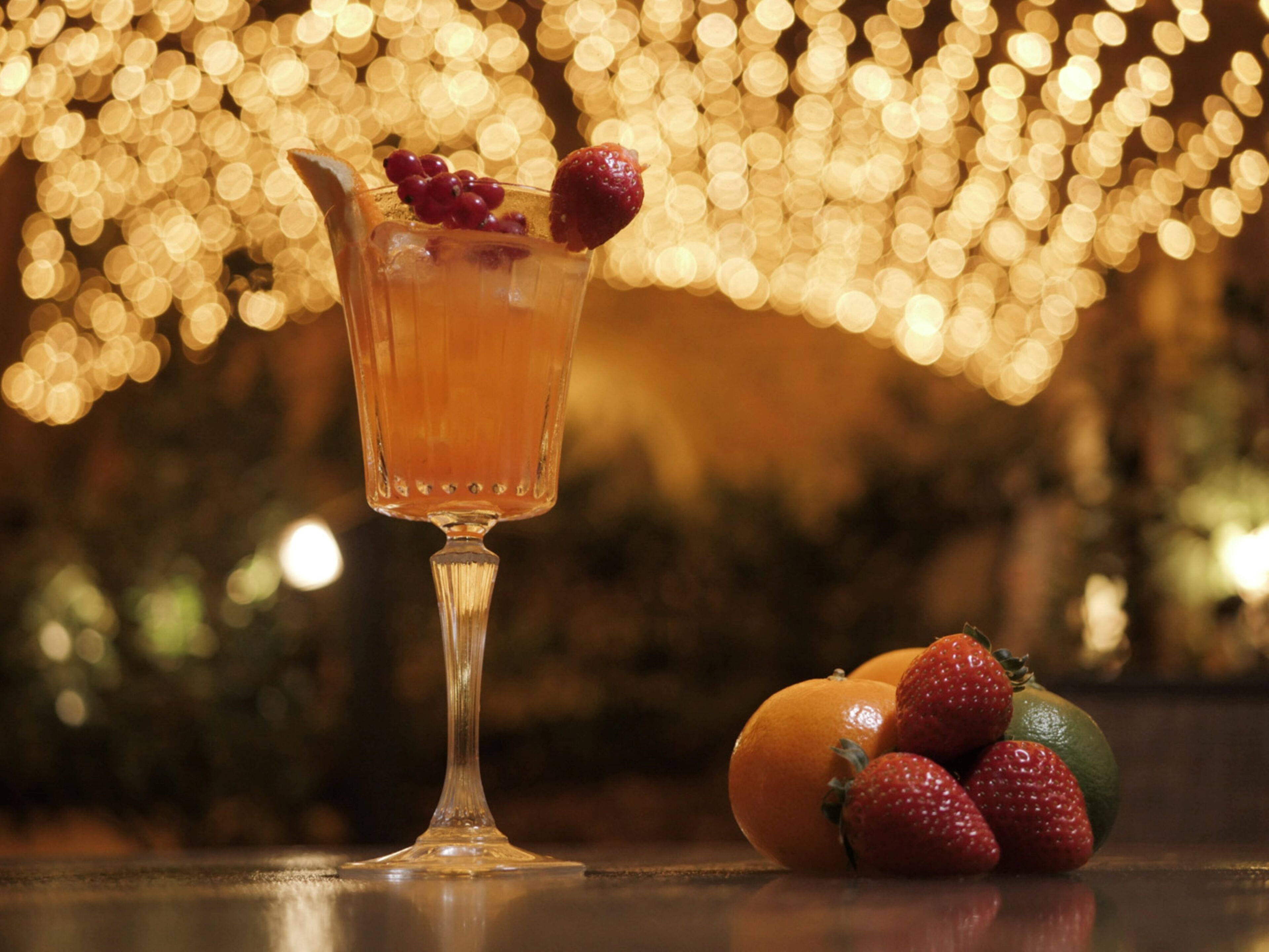 A cocktail glass with a fruit garnish on a reflective surface, accompanied by oranges and strawberries.