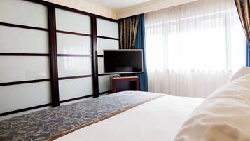 Une chambre d’hôtel avec un lit, un téléviseur posé sur un meuble, un tapis à motifs et une fenêtre avec des rideaux.