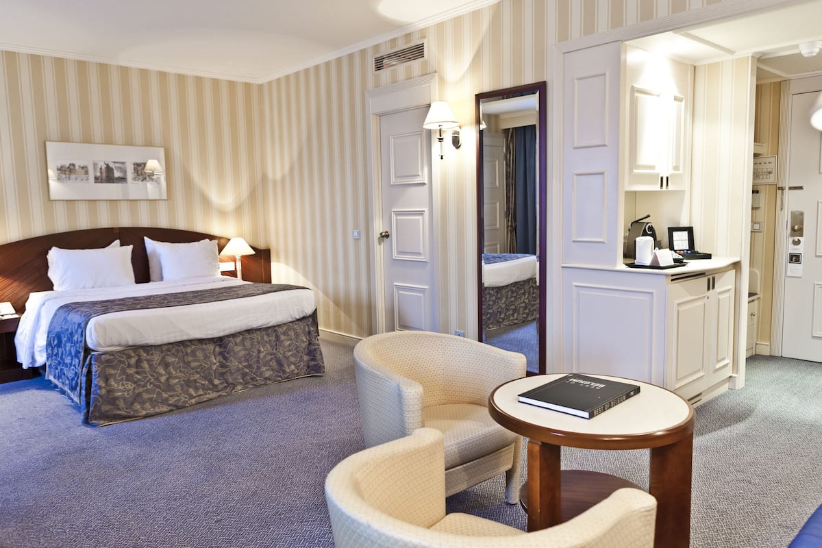 A hotel room with a bed, a chair, a small table, a mirror, and a kitchenette.