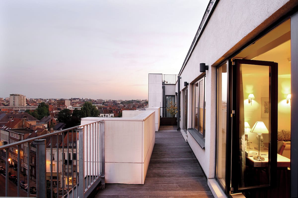 A modern hotel balcony with a wooden floor, a seating area with a table and chairs, and a view of the cityscape.