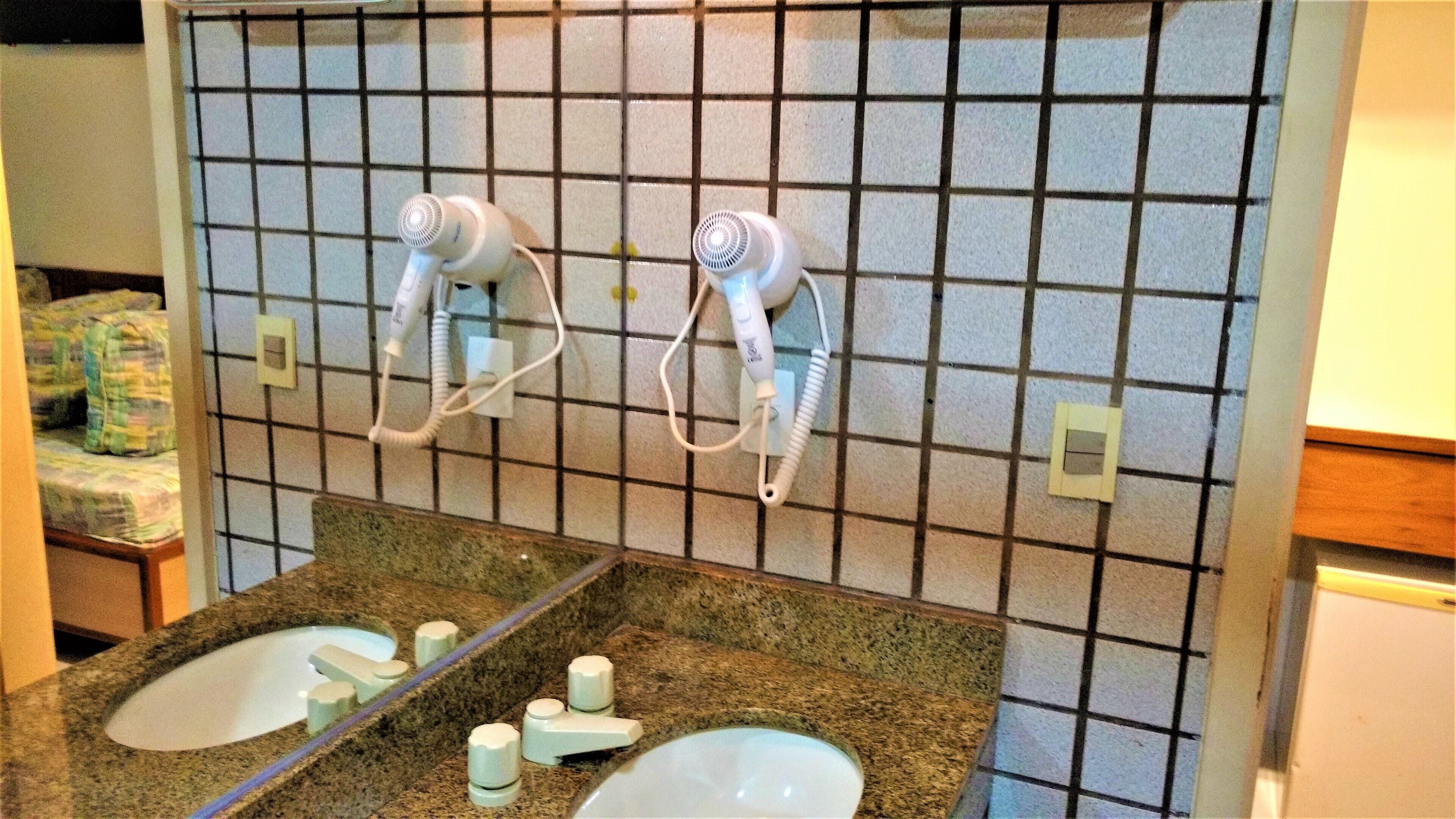 Baño con salpicadero de azulejos, dos secadores de pelo montados en la pared y encimera de granito con un espejo y dos lavamanos.