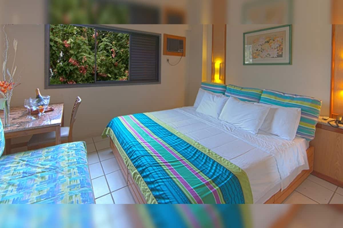 A bedroom with a bed, a bedside table, a chair, a window with a view of greenery, and a wall-mounted air conditioning unit.