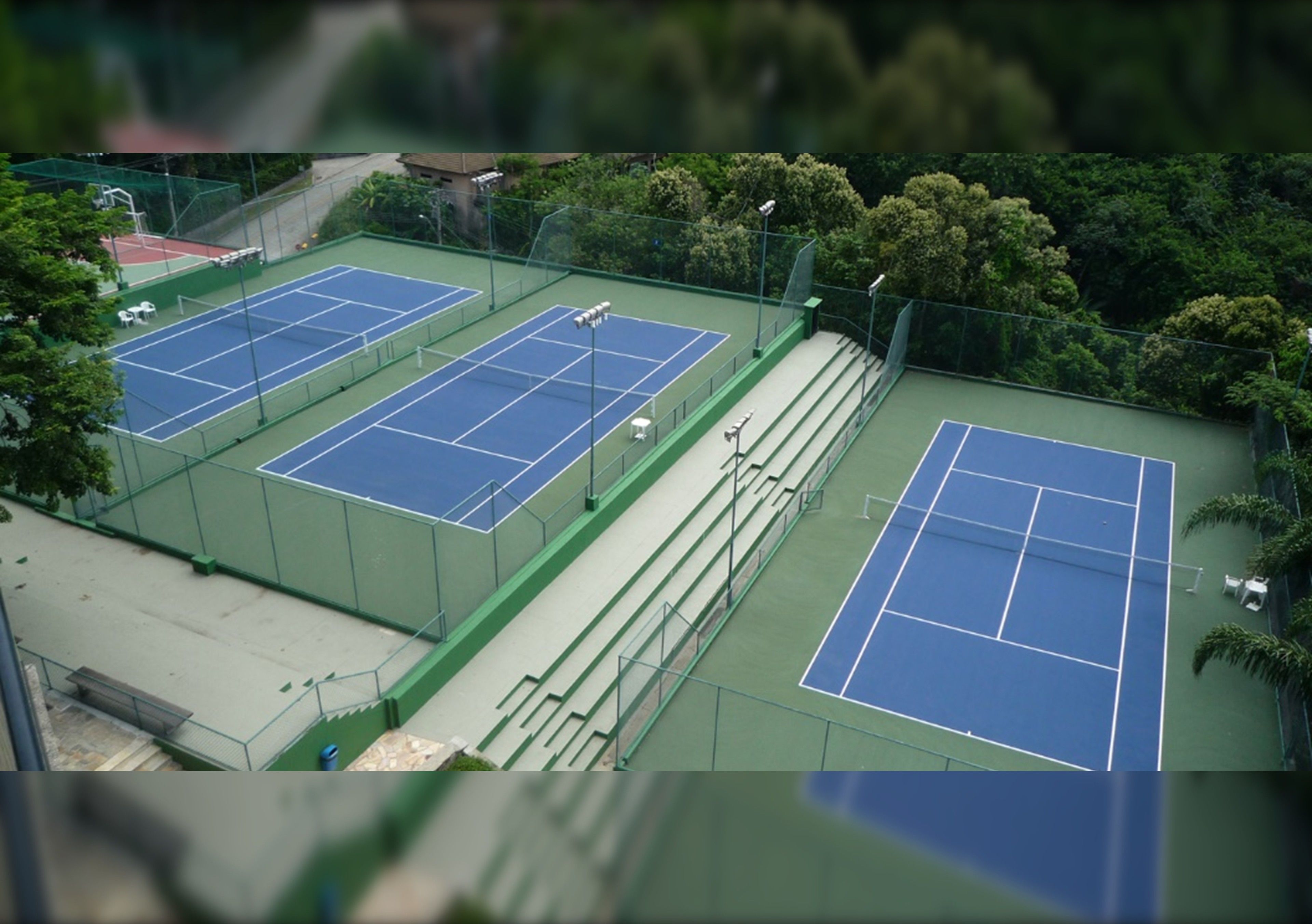 An aerial view of a tennis court complex with multiple courts, surrounded by a green fence and trees.