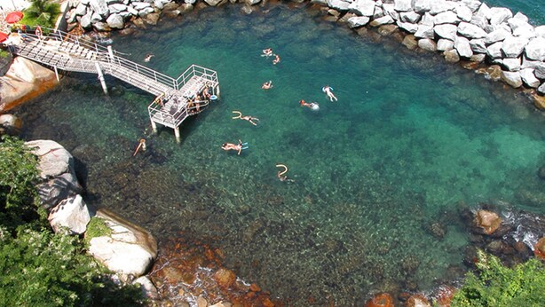 Um corpo de água límpida rodeado por pedras, uma ponte de madeira e uma pequena plataforma onde pessoas nadam.