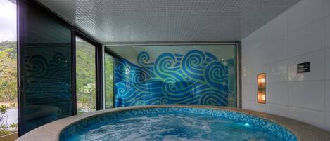 A modern indoor swimming pool with a glass wall featuring blue wave patterns, a tiled ceiling, and a view of greenery outside.