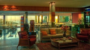 Um hall de hotel com balcão de recepção, área de estar com sofás e poltronas, um grande pilar e vista para o oceano através de amplas janelas.