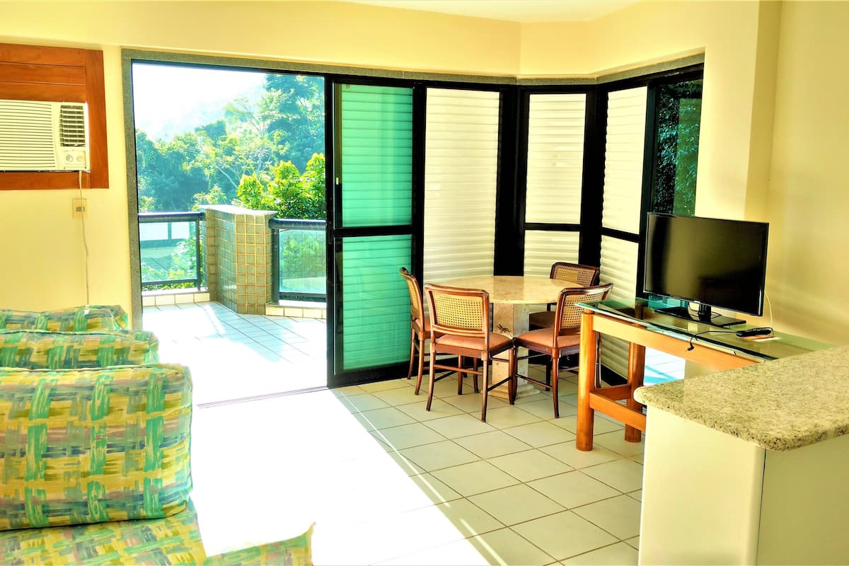 A room with a TV, a table, and two chairs. There is a window with blinds and a view of greenery outside.