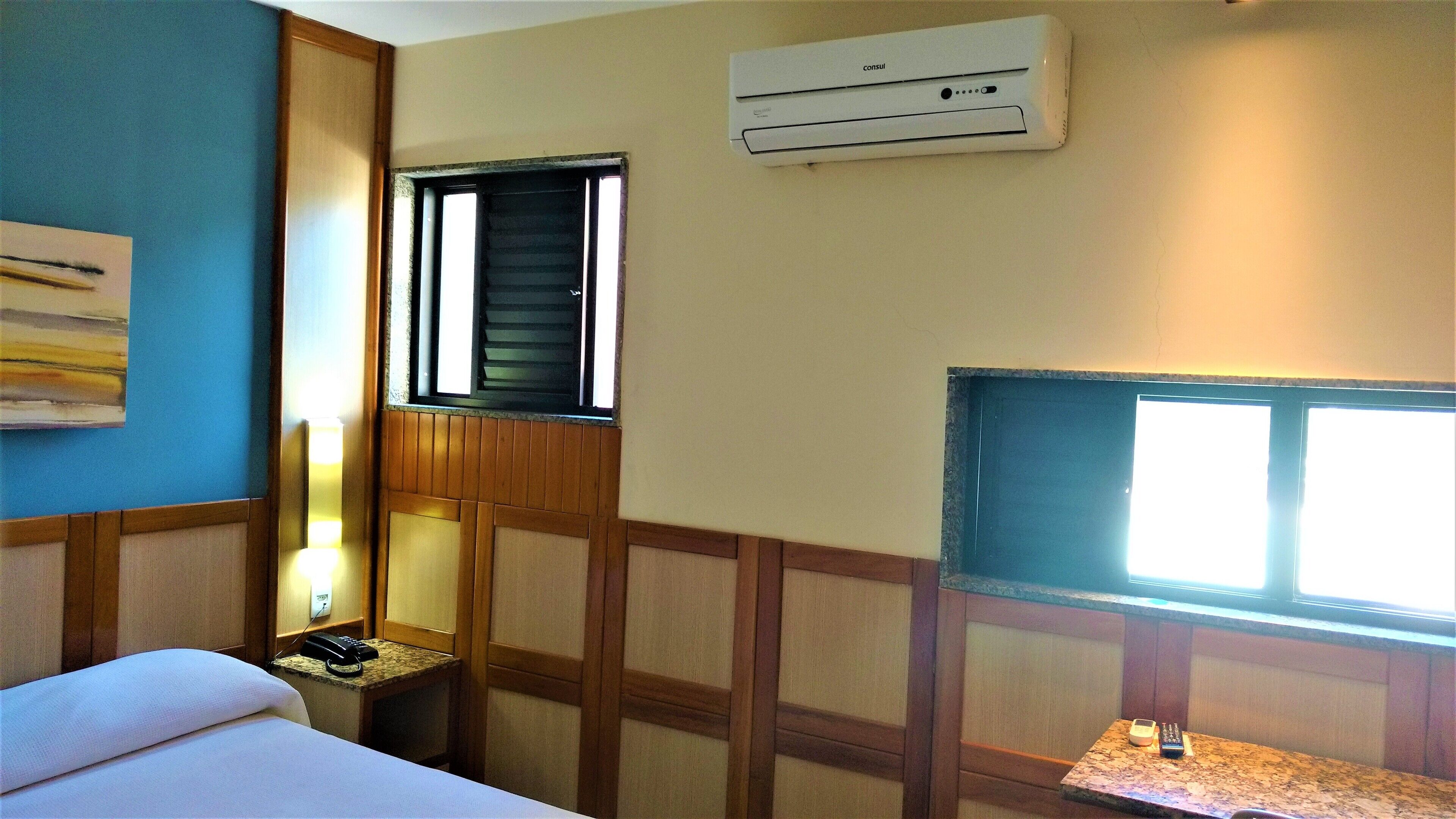 A hotel room with a bed, a desk with a telephone, a window with blinds, and an air conditioning unit on the wall.