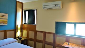 A hotel room with a bed, a desk with a telephone, a window with blinds, and an air conditioning unit on the wall.