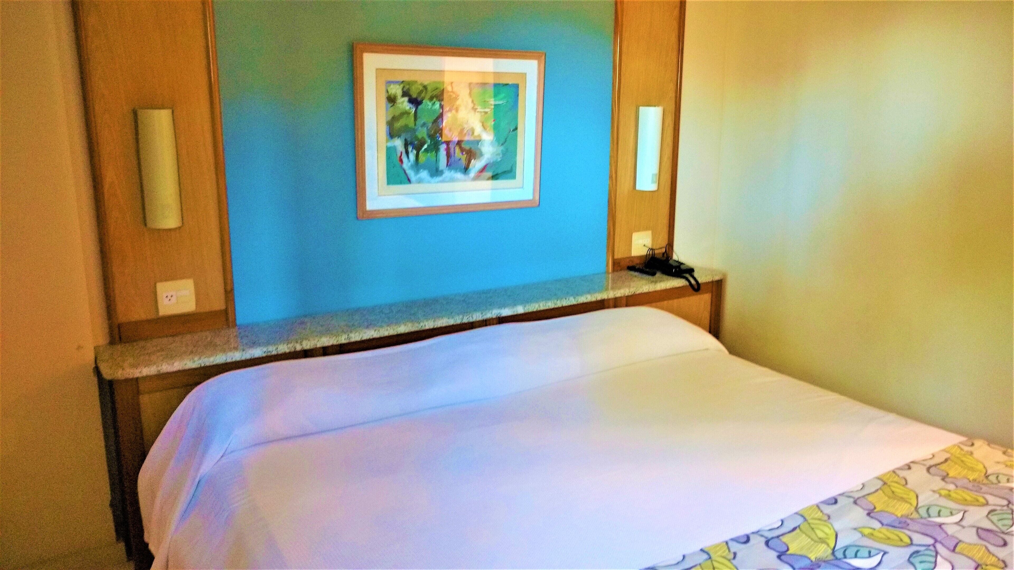 A hotel room with a bed, a framed artwork on the wall, and a nightstand with a phone.