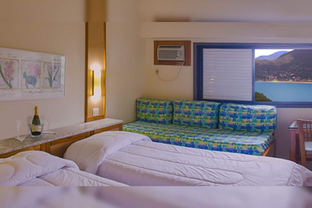A hotel room with a bed, a small table with a bottle and glass, a sofa with a colorful cushion, and a window with a view of a lake and mountains.