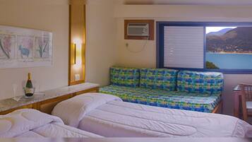 A hotel room with a bed, a small table with a bottle and glass, a sofa with a colorful cushion, and a window with a view of a lake and mountains.