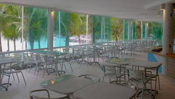 Uma ampla área de refeições interna com grandes janelas que dão para uma praia e palmeiras. Mesas e cadeiras dispostas para refeições.