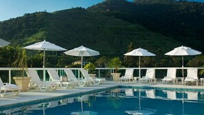 Área de piscina com espreguiçadeiras e guarda-sóis, cercada por vegetação exuberante e colinas.