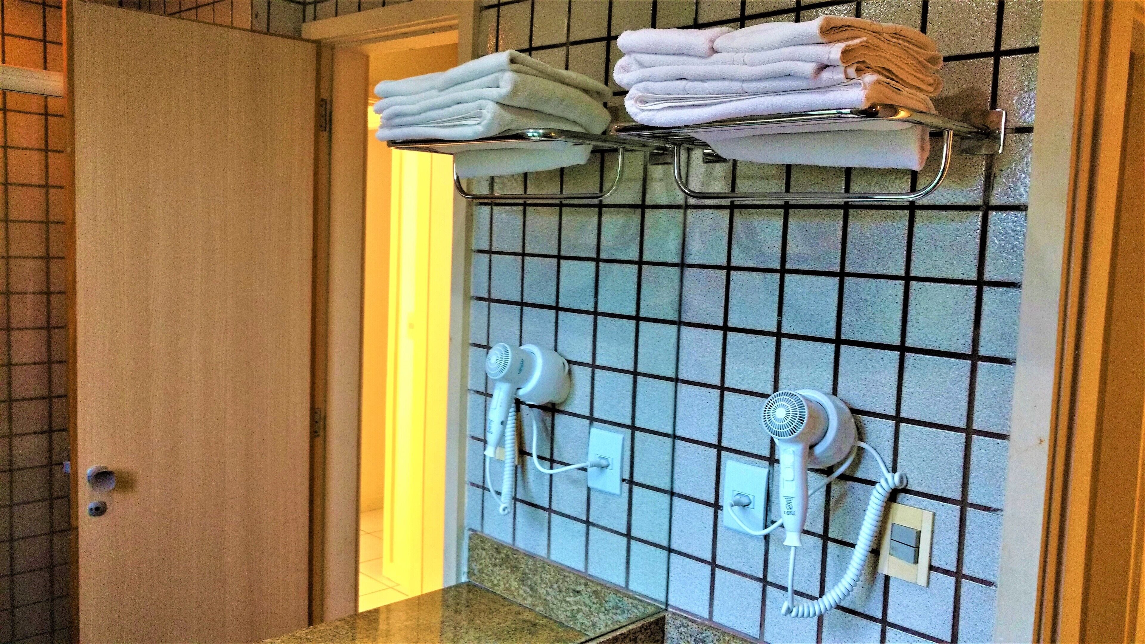 A bathroom with a tiled wall, a towel rack with folded towels, and two hair dryers mounted on the wall.