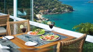 Uma mesa com pratos de comida e um copo de vinho tinto, com vista para a costa, casas e vegetação.