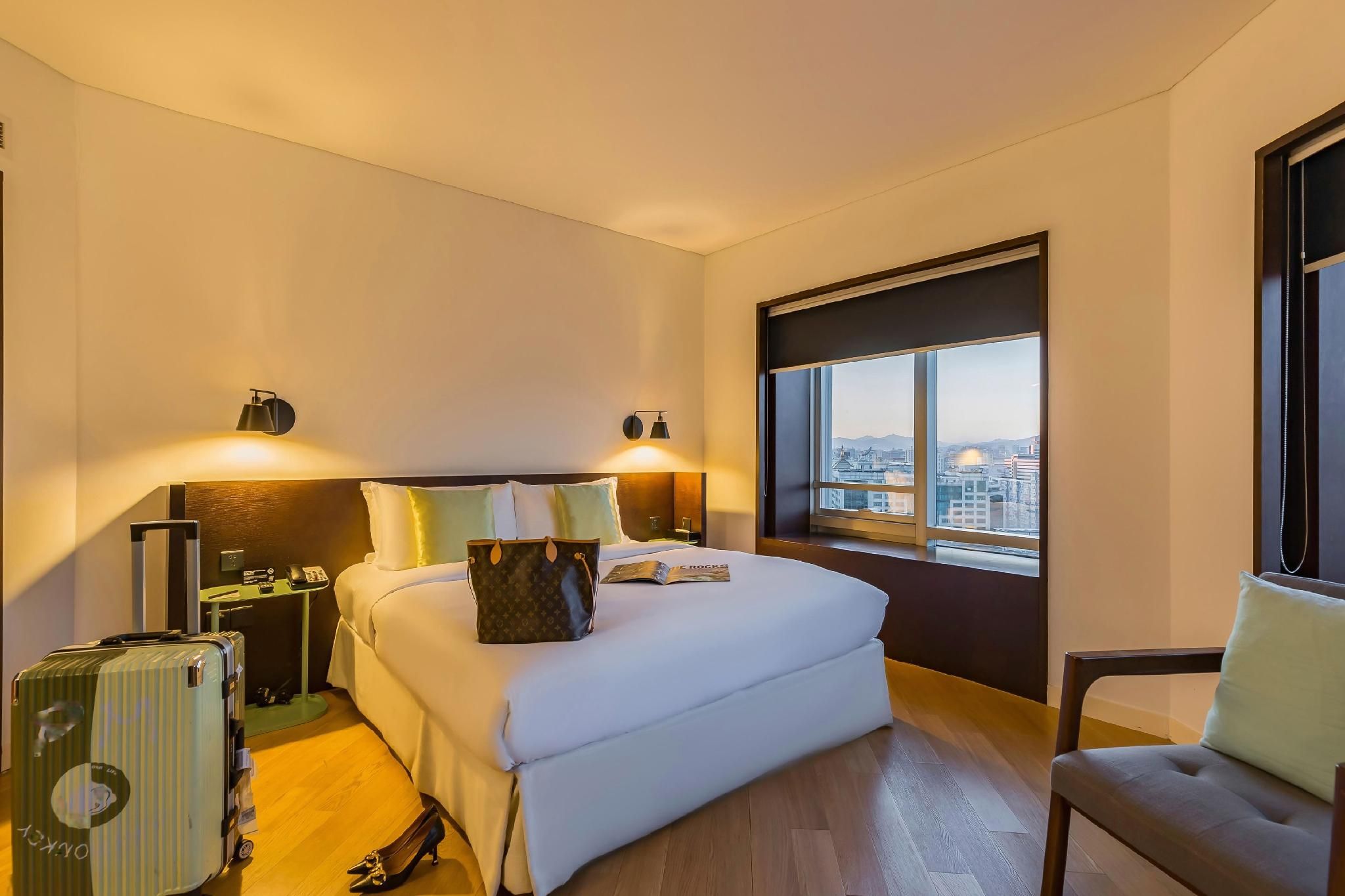 A hotel room with a bed, bedside lamps, a suitcase, a chair, and a window with a city view.