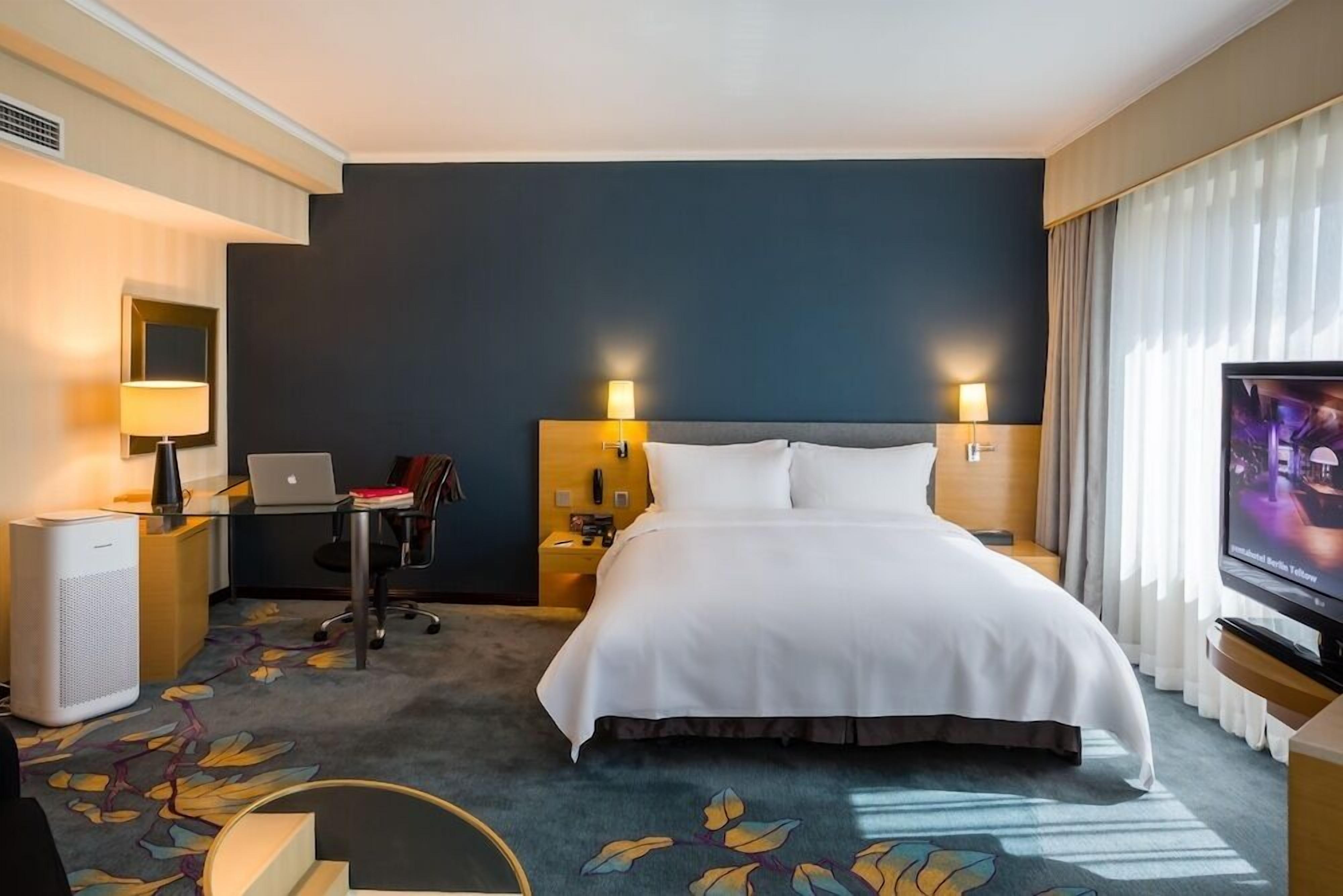 Ein Hotelzimmer mit Bett, Schreibtisch, Stuhl, Fernseher und einem großen Fenster mit Vorhängen.