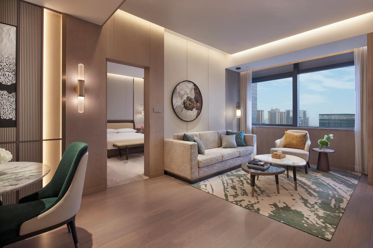 A modern hotel room with a sofa, armchair, and coffee table. A bed is visible in the background, and there is a large window with a city view.