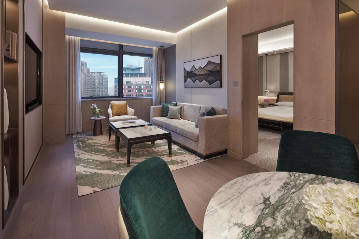 A modern hotel room with a sofa, a coffee table, a dining table, and a view of the cityscape through large windows.