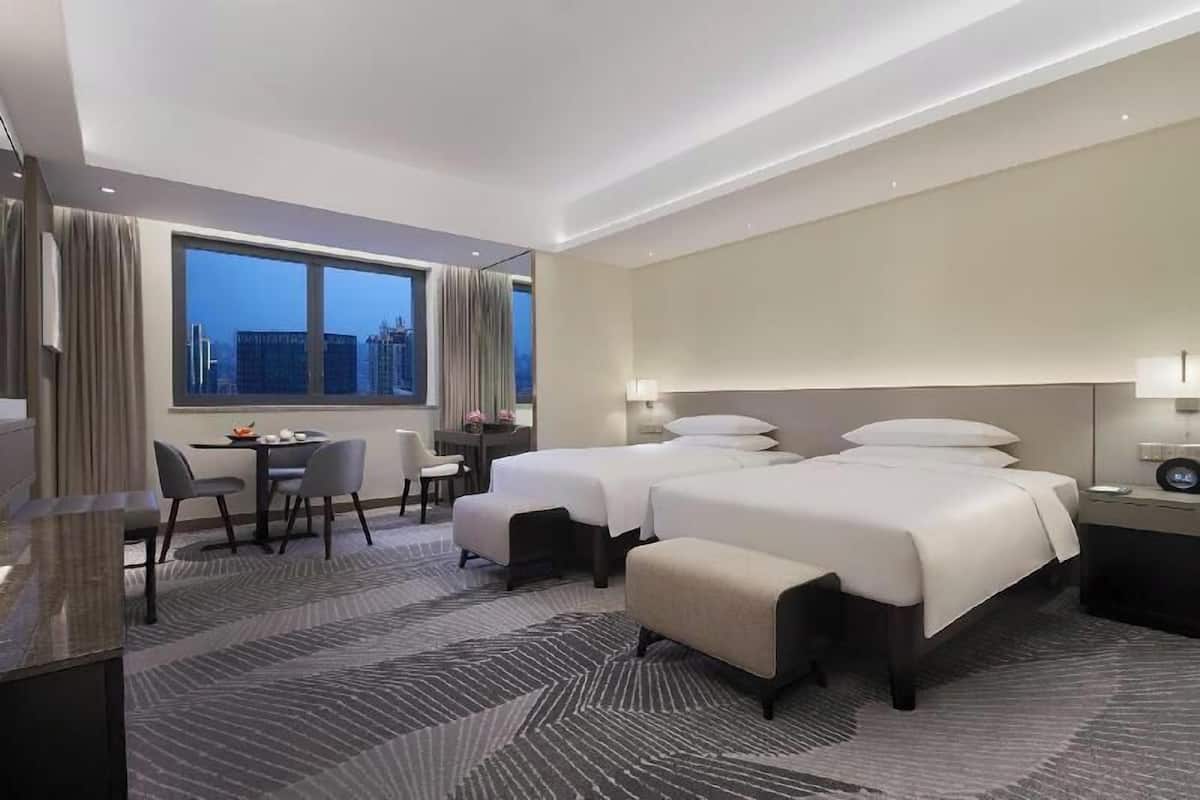 A hotel room with two beds, a desk, a chair, and a view of the cityscape.