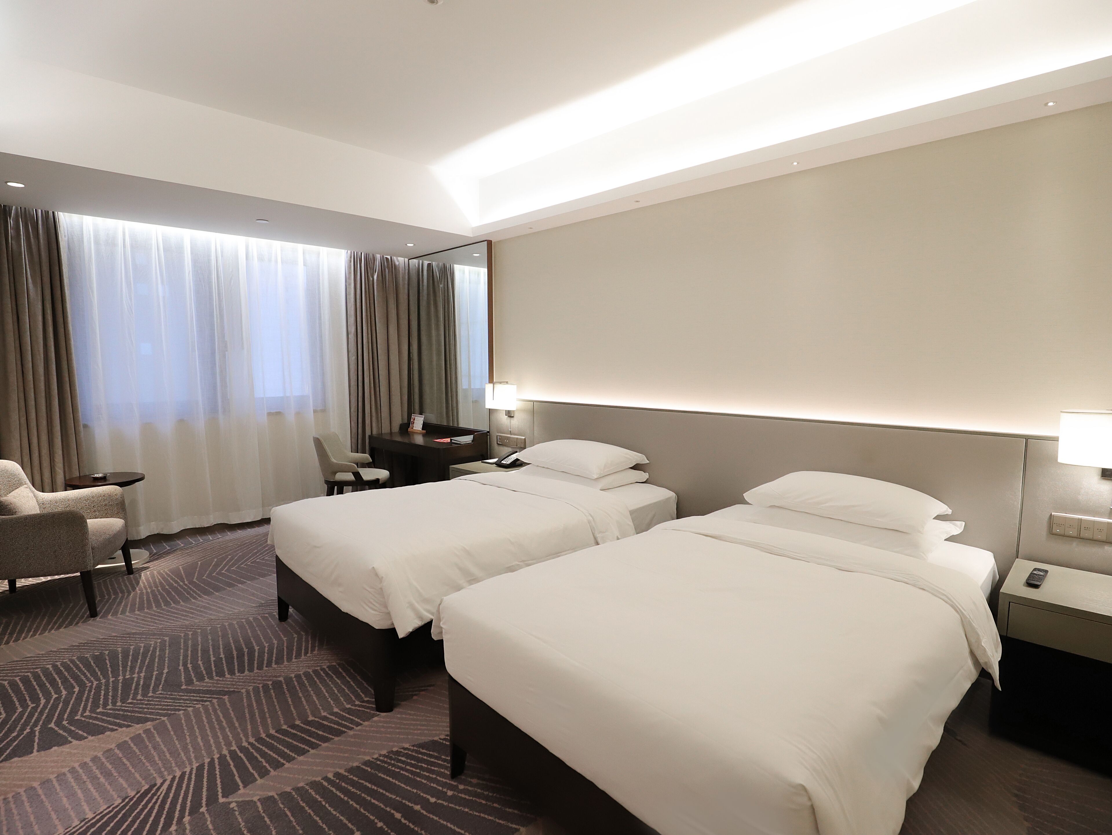 Ein Hotelzimmer mit zwei Betten, einem Schreibtisch, einem Stuhl und einem großen Fenster mit Vorhängen.