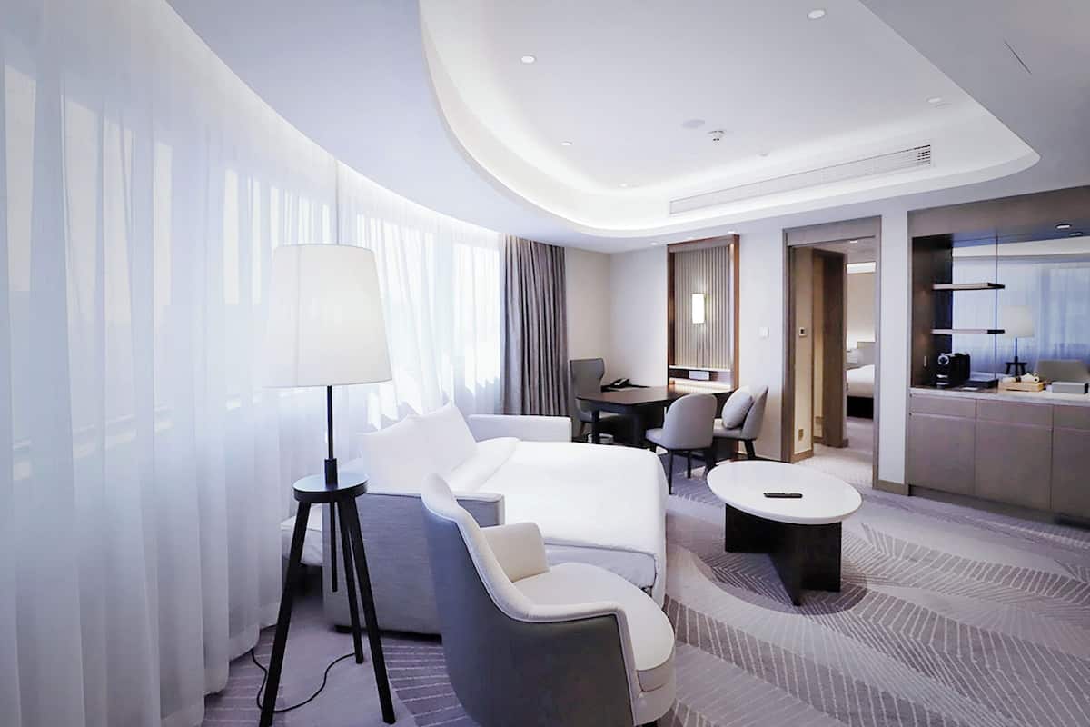 A modern hotel room with a large bed, a seating area with a sofa and chairs, a dining table, and a large window with sheer curtains.