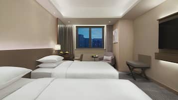Habitación de hotel con dos camas, un escritorio, una silla, una mesita con frutero, una ventana con vista a la ciudad y un televisor montado en la pared.