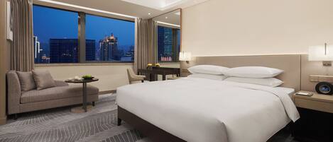 A hotel room with a large bed, a desk, a chair, and a view of the cityscape at night.