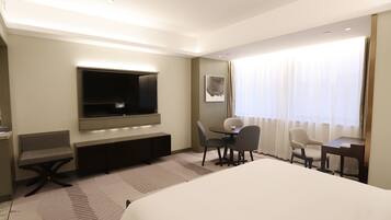 Une chambre d’hôtel avec un grand lit, un bureau avec deux chaises, un téléviseur fixé au mur et une fenêtre avec des rideaux.