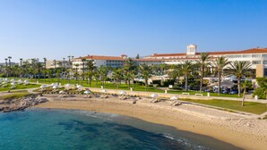 A resort with a beach, palm trees, and a large building with a tower. - Olympic Lagoon Resort - Paphos - All inclusive (Paphos)