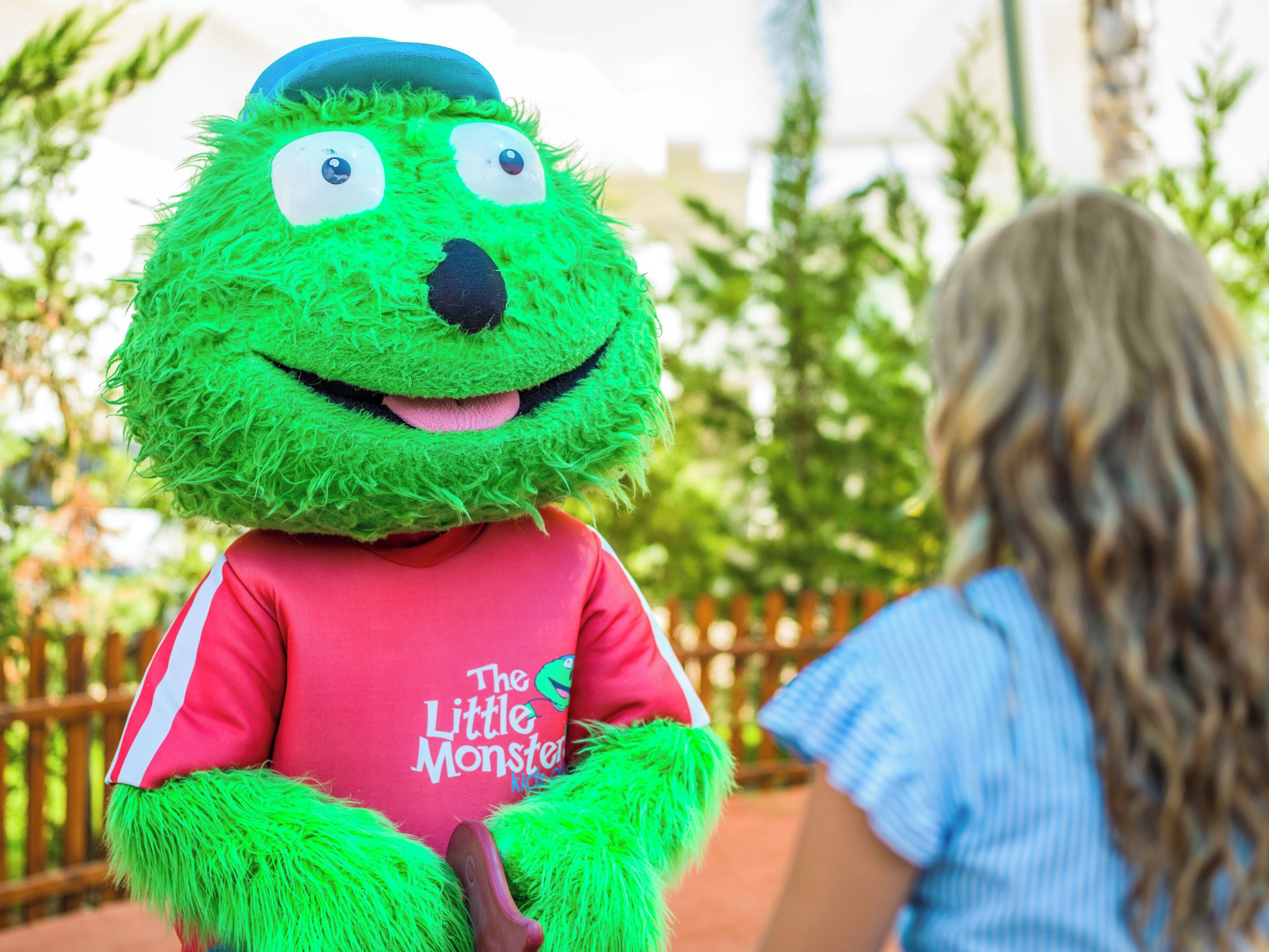 Une mascotte portant un t-shirt rouge avec l’inscription « The Little Monster Kids », debout à l’extérieur, avec de la verdure en arrière-plan.