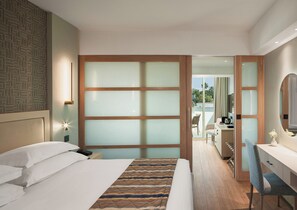 A hotel room with a bed, a desk, a chair, a mirror, and a view of the outdoors. - Olympic Lagoon Resort - Paphos (Paphos)