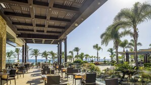 An outdoor dining area with tables and chairs, palm trees, and a view of the ocean. - Olympic Lagoon Resort - Paphos - All inclusive (Paphos)