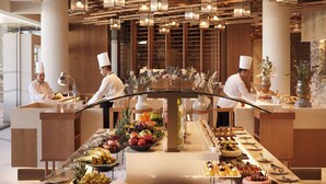 A modern hotel lobby with a central buffet table, wooden ceiling design, and staff in chef uniforms. - Olympic Lagoon Resort - Paphos - All inclusive (Paphos)