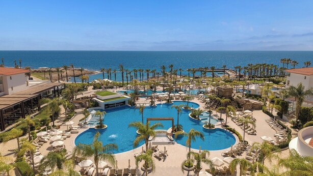 A resort with a central pool, palm trees, and a view of the ocean. - Olympic Lagoon Resort - Paphos - All inclusive (Paphos)
