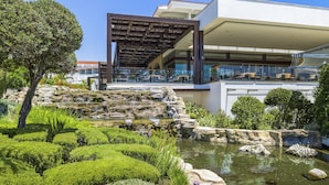 A modern building with a rooftop terrace, a landscaped garden with a pond, and a rocky waterfall feature. - Olympic Lagoon Resort - Paphos - All inclusive (Paphos)