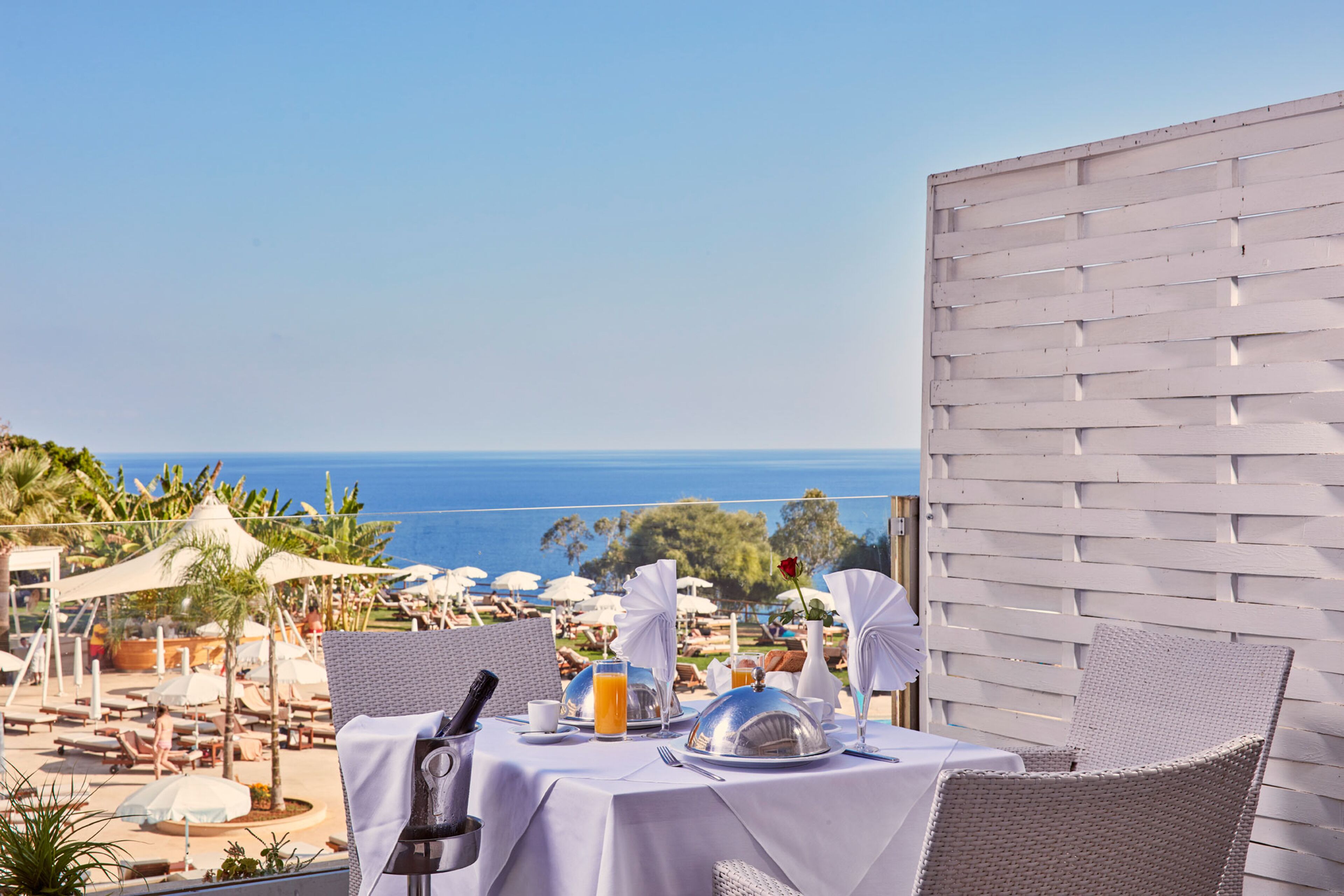A table set for breakfast with a view of the ocean, white wicker chairs, and umbrellas in the background.