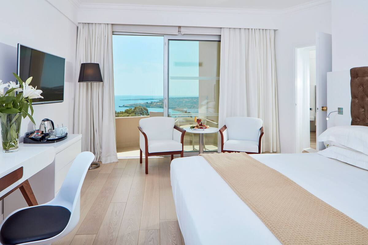 Une chambre d’hôtel avec un lit, deux fauteuils blancs, une petite table, un téléviseur et une vue sur la mer.