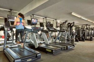Uma academia com esteiras, aparelhos de ginástica e uma televisão exibindo um programa de exercícios.