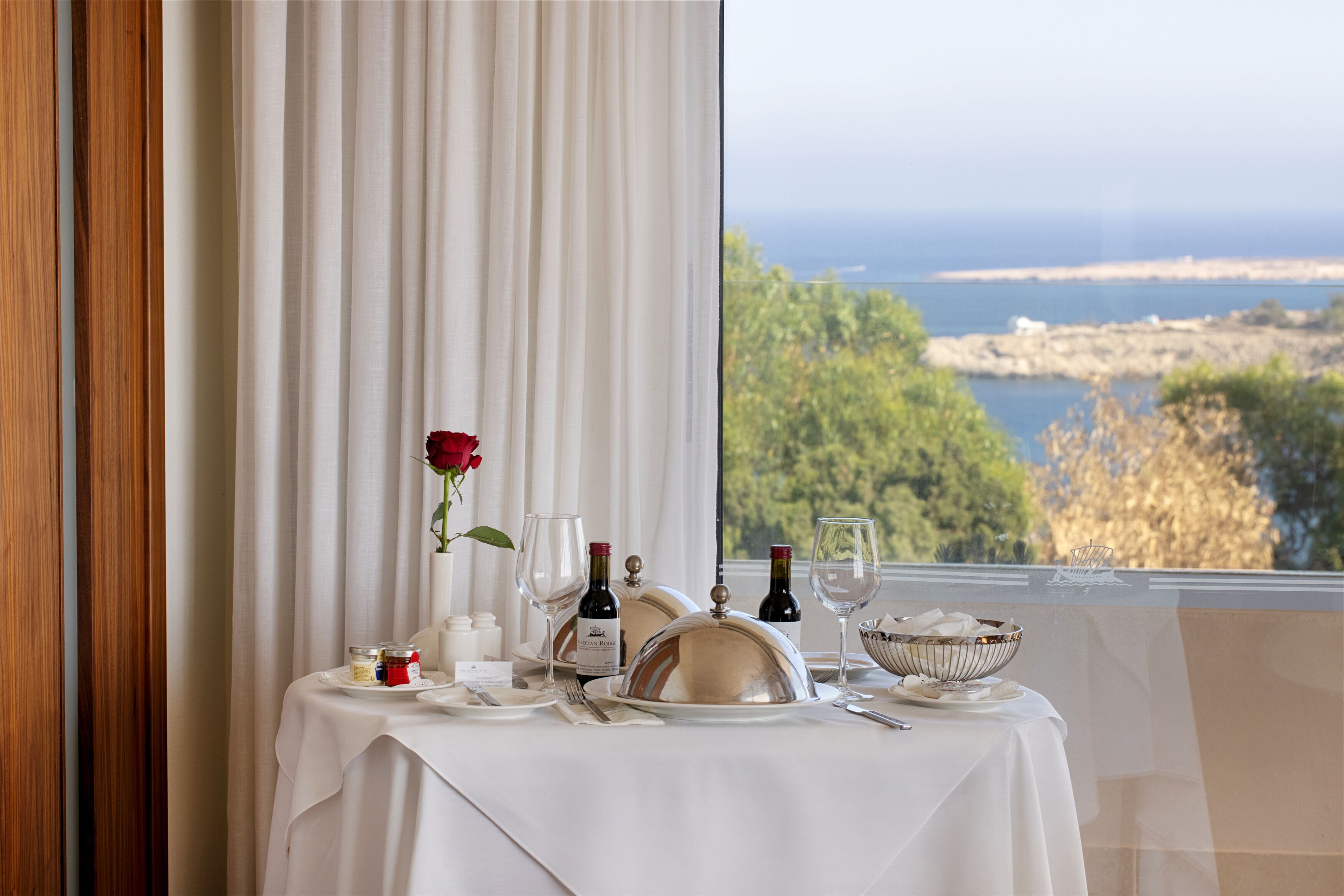 A table set for two with wine glasses, a rose, and a view of the sea.
