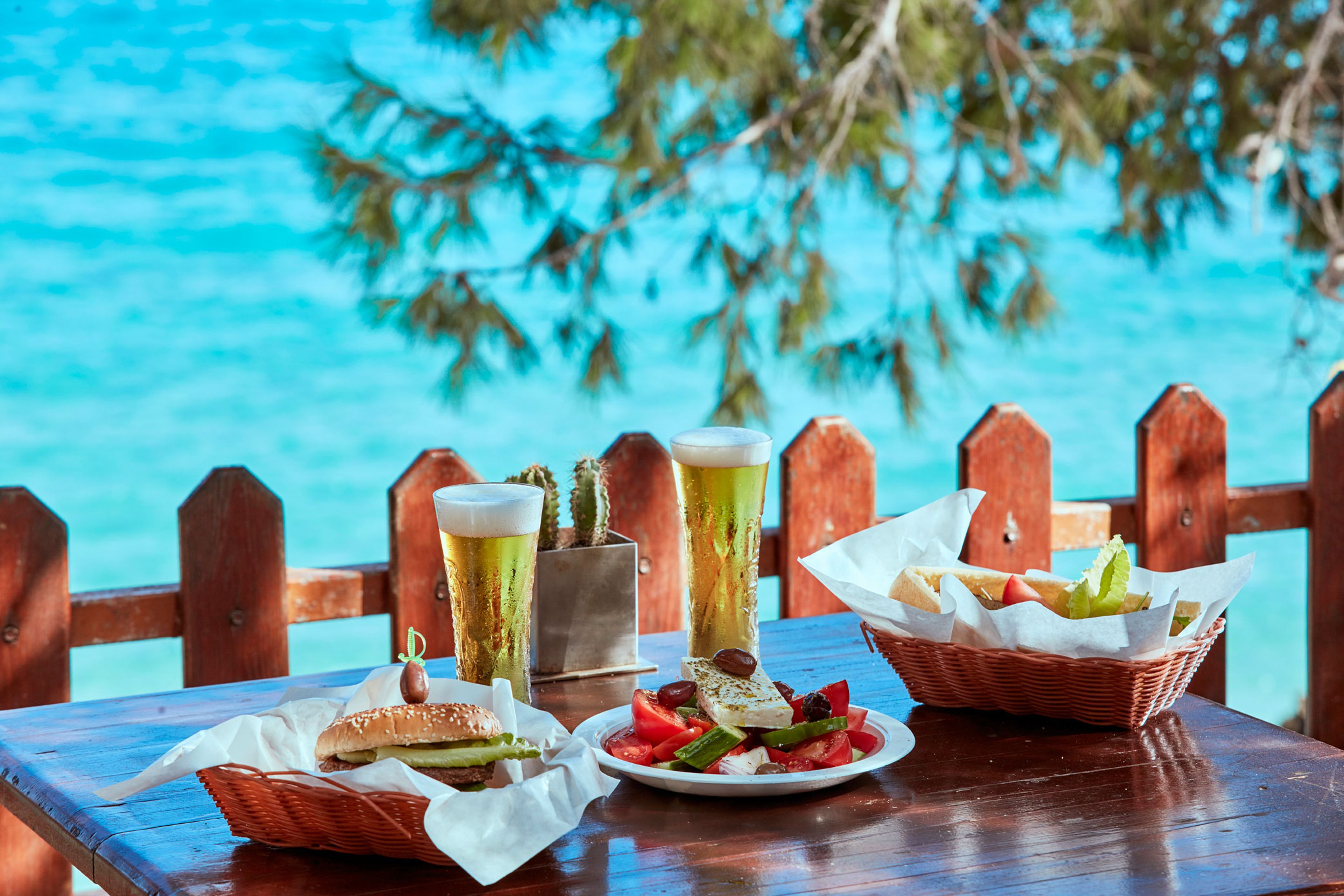 A wooden table with a plate of food, a basket of bread, and two tall glasses of beer.