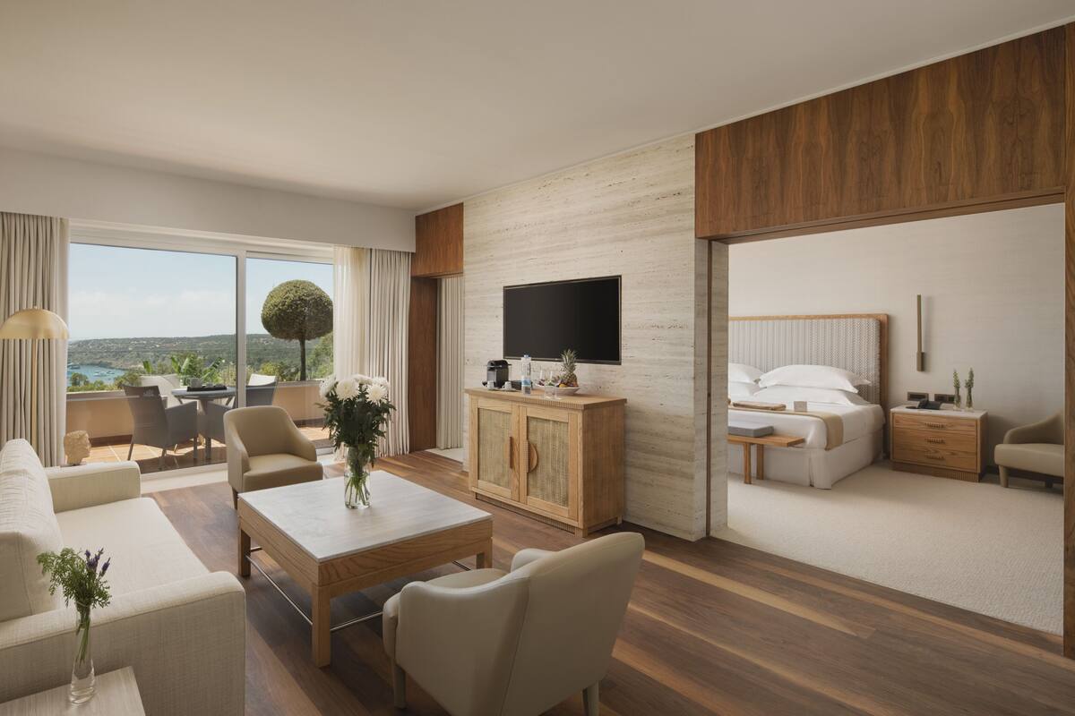 Une chambre d’hôtel moderne dotée d’une grande fenêtre, d’un téléviseur à écran plat, d’une armoire en bois, d’un canapé, d’une table à manger et d’un lit avec une table de chevet.