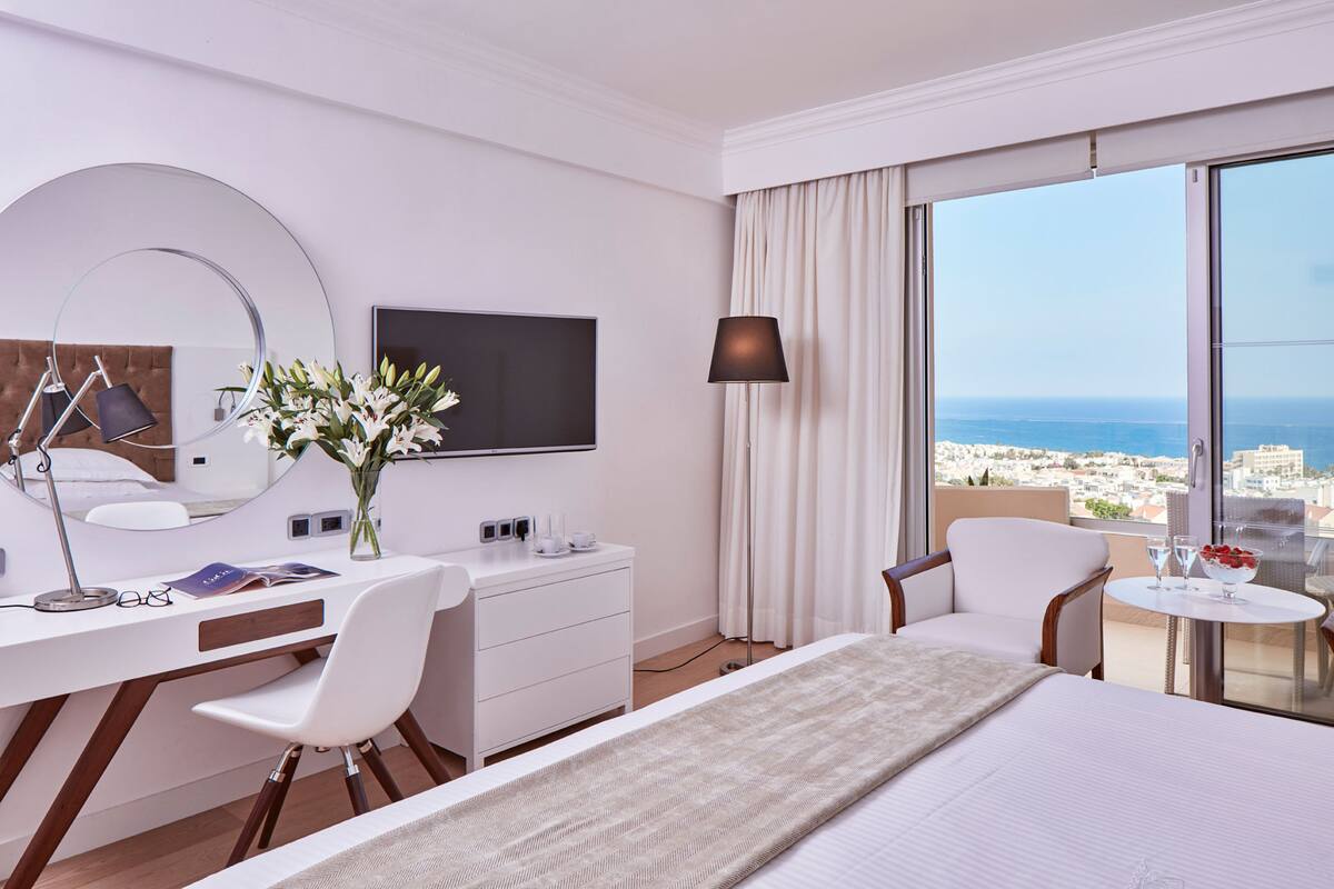 Une chambre d’hôtel moderne équipée d’un lit, d’un bureau, de chaises, d’une télévision et d’un balcon avec vue sur la mer.