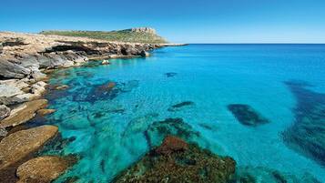Paisaje costero con agua turquesa cristalina, costa rocosa y una colina distante.