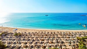 A beach with rows of sun loungers and thatched umbrellas, a clear blue sea, and a few boats in the distance. - Grecian Park Hotel (Paralimni)