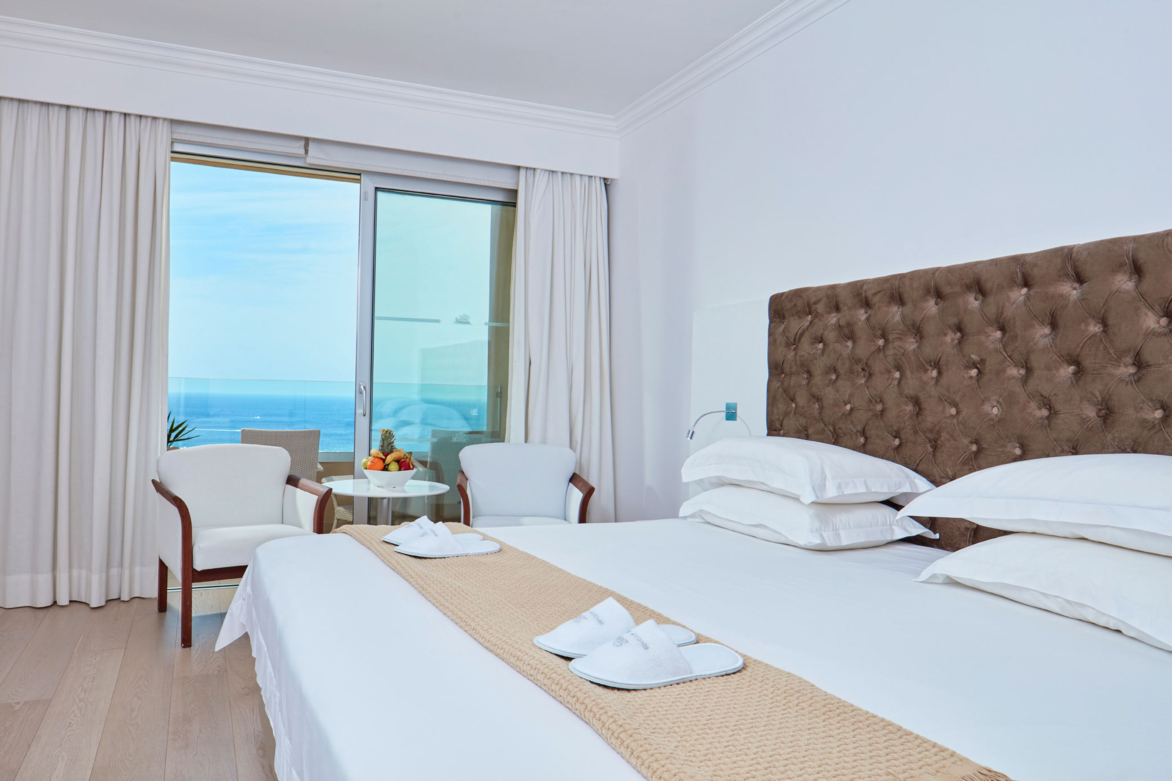 A hotel room with a large bed, a tufted headboard, a view of the ocean, a small round table with fruit, and a chair.