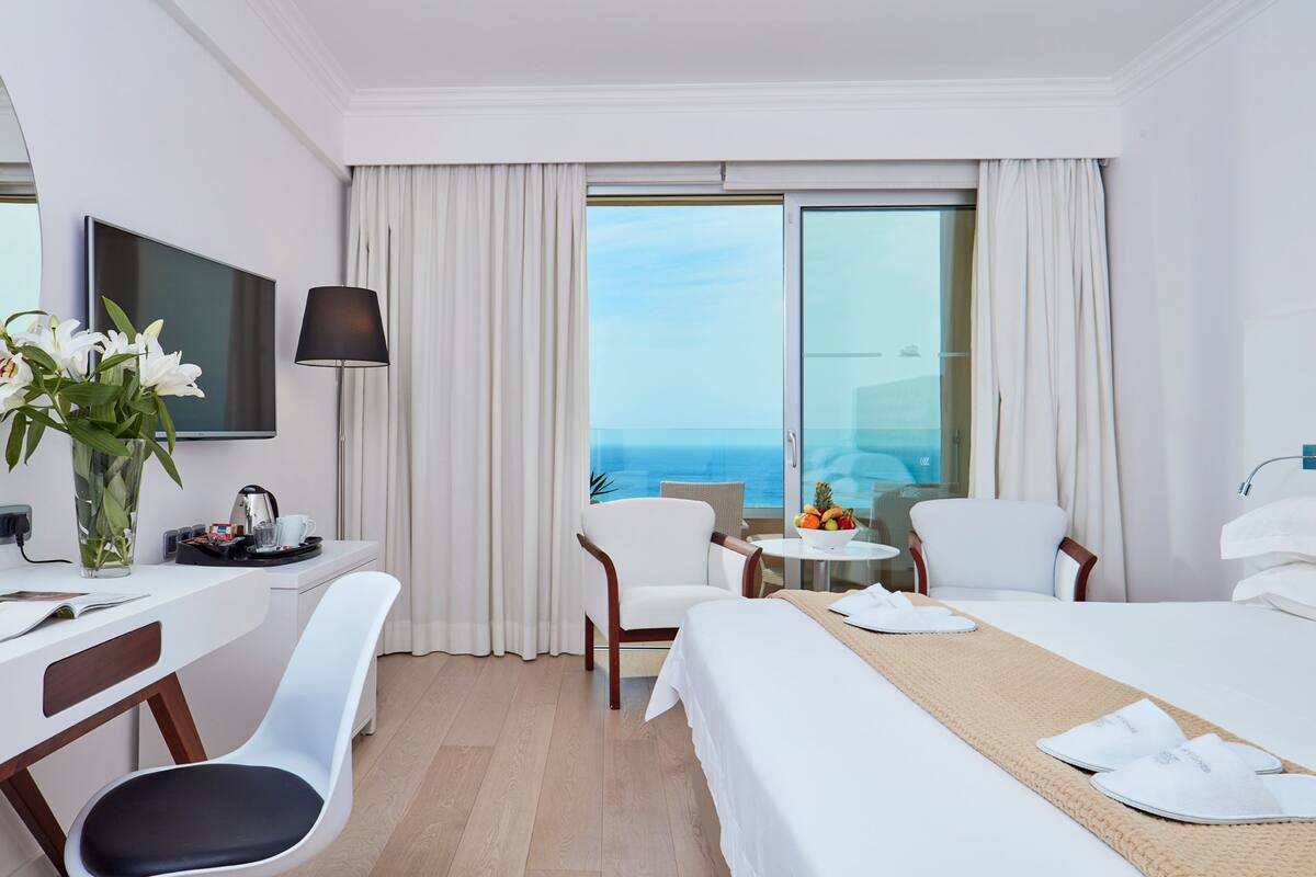 Une chambre d’hôtel avec un lit, deux chaises, un bureau avec un ordinateur portable, une télévision et une vue sur l’océan.