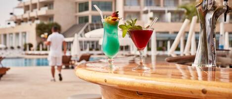 A bar with two cocktails on a wooden bar counter, a pool, and a building in the background.
